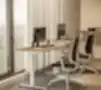 Modernios biuro erdvės su dviem ergonomiškais reguliuojamo aukščio stalais prie didelio lango, aprūpintomis juodomis tinklinėmis kėdėmis, kompiuterių monitoriais ir mediniais stalviršiais, apšviestomis natūralia šviesa.