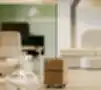 Moderni biuro kampelis su balta ergonomiška biuro kėde, reguliuojamo aukščio stalu su kompiuterio monitoriumi ir tamsiai žalia privatumo pertvara, mediniu mobiliu stalčiu ant ratukų, šviesiomis medinėmis grindimis, baltu moduliuotu sofa fone ir dideliu kreminiu pakabinamuoju šviestuvu ant lubų.