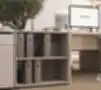 Žema atvira biuro lentyna smėlio spalvos, su dviem lentynomis segtuvams laikyti, šalia darbo stalo su kompiuteriu ir alyvmedžio vazonu.