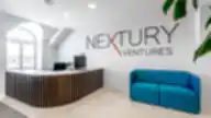 Modernus „Nextury Ventures“ biuro registratūros priimamasis su medine lenkta stalo apdaila, baltais sienų paviršiais, mėlyna sofa ir įmonės logotipu ant sienos.