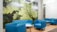 Keletas ryškiai mėlynų, vertikaliai siūtais raštais dekoruotų fotelių ir mažų baltų apvalių stalelių stovi šviesaus medžio grindyse prieš didelį žalialapių ąžuolo lapų sieninį piešinį. Prie sėdėjimo zonos stovi lygus betono stulpas, o iš baltos grotelėmis dengtos lubos kabantys kvadratiniai LED šviestuvai skleidžia tolygų apšvietimą.