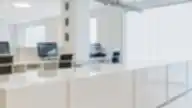 Moderni, balta Clinic DPC dantų priežiūros centro registratūra su kompiuteriais ir kėdėmis už stiklinės sienos su įmonės logotipu.