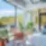 Šviesi SHED Co-living Vilnius poilsio erdvė su vazoniniais augalais ir pastelinėmis kėdėmis, dideliais langais ir išėjimu į terasą su vaizdu į mišką.