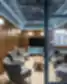 Stiklinių pertvarų atskirta susitikimų poilsio zona verslo centre Business Garden Lounge su pilkomis liūtėtomis fotelėmis aplink medinį kavos stalelį, turkio spalvos medijos spintele po kabinamu ekranu, šiltomis medinėmis panelėmis, sferiniais sieniniais šviestuvais ir metalinio tinklelio lubomis.
