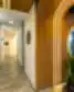 Neries odontologijos klinikos koridorius su baltomis durimis, šviesiomis grindimis ir moderniu apšvietimu, dešinėje matoma medinė sienelė su LED apšvietimu ir augalais.