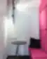 Mažas poilsio kampelis su ryškiai rožiniu minkštasuoliu, juodu staliuku, juoda dekoratyvine pagalve ir sieniniu apšvietimu.