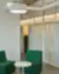 Minimalistinė biuro poilsio zona su dviem žaliomis kėdėmis, baltu apvaliu staliuku, medinėmis pertvaromis ir matomomis ventiliacijos sistemomis.